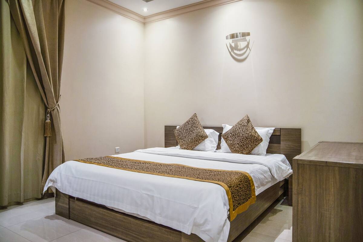 Una cama bien hecha con una colcha blanca y marrón, dos almohadones color leopardo y un cabecero de madera.