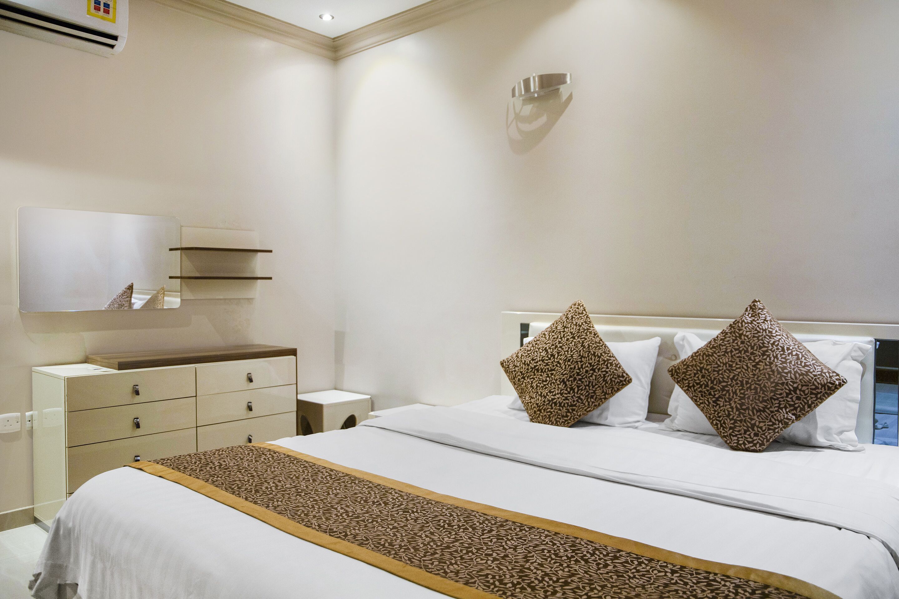 Habitación de hotel con cama, cómoda, mesita de noche, espejo y estante en la pared.
