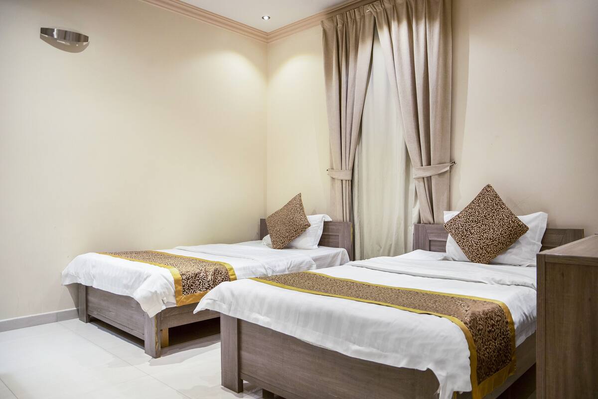 Habitación de hotel con dos camas individuales, cada una con una colcha blanca y un corredor estampado. La habitación tiene una paleta de colores neutra, con paredes color beige y un cabecero de madera.