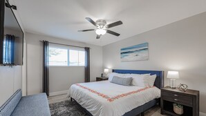 Un dormitorio con cama, mesita de noche, banco, ventilador de techo, ventana con cortinas y un cuadro en la pared.