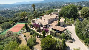Une villa dotée d’un court de tennis, entourée d’une végétation luxuriante et avec des montagnes en toile de fond.