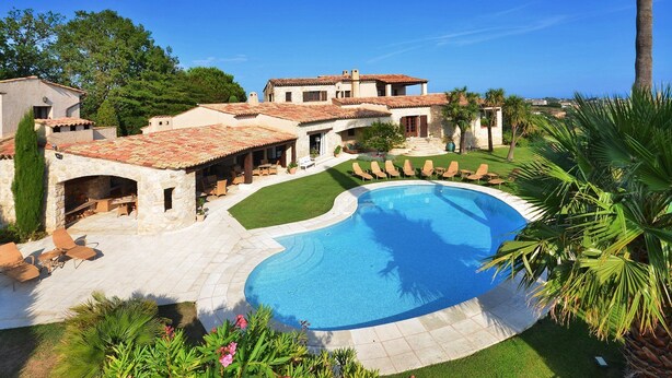 A large, curved swimming pool surrounded by a stone patio, with outdoor seating and palm trees.