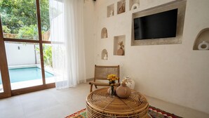 A room with a large window, a wicker chair, a round table with a vase of flowers, and a flat-screen TV mounted on the wall. - Villa Milos (Canggu)