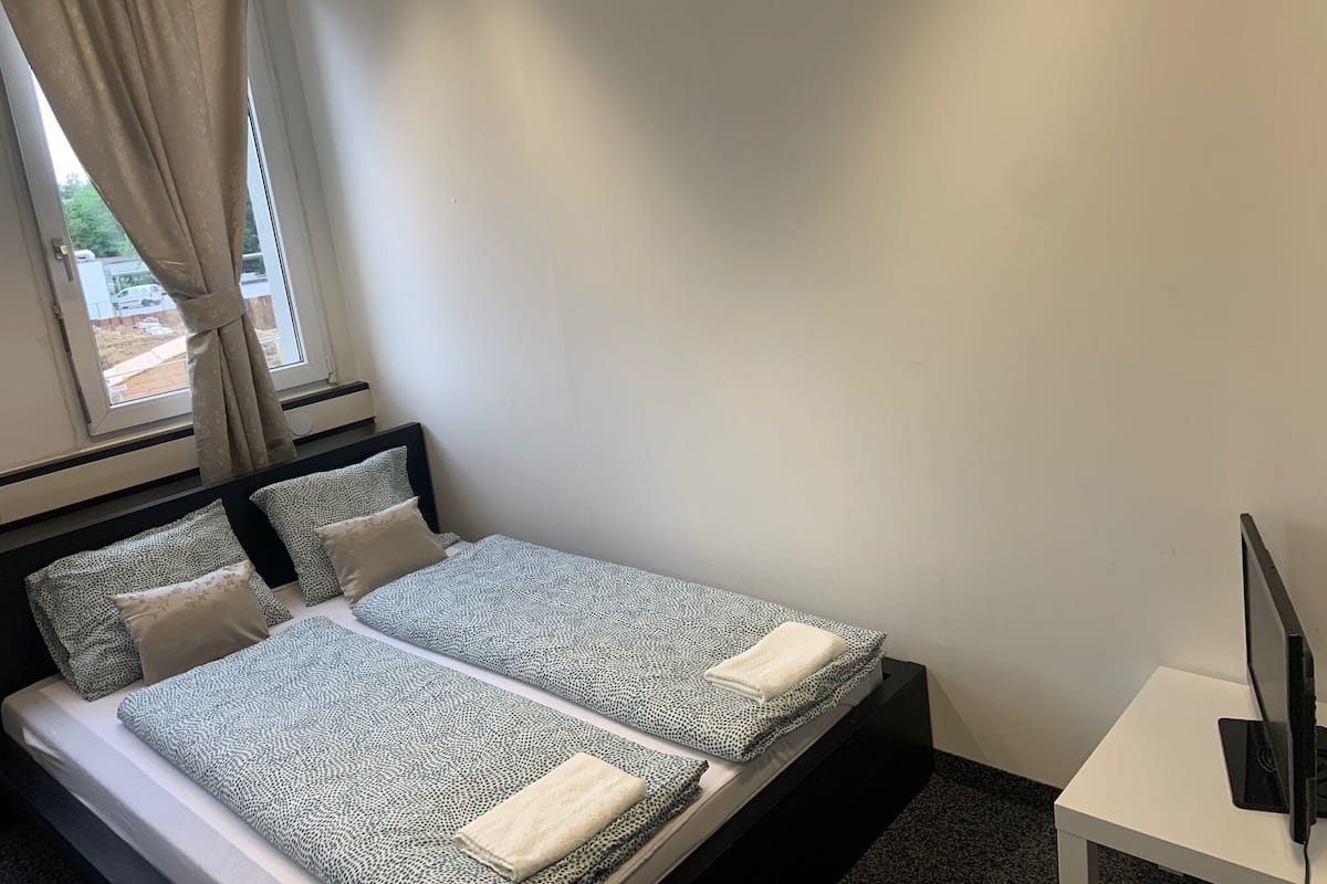 A double bed with patterned bedding, a bedside table with a flat-screen TV, and a window with curtains.