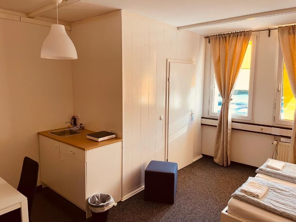 Ein kleines Hotelzimmer mit Küchenzeile, einem kleinen Hocker, einem Bett mit Handtüchern und einem Fenster mit Vorhängen.