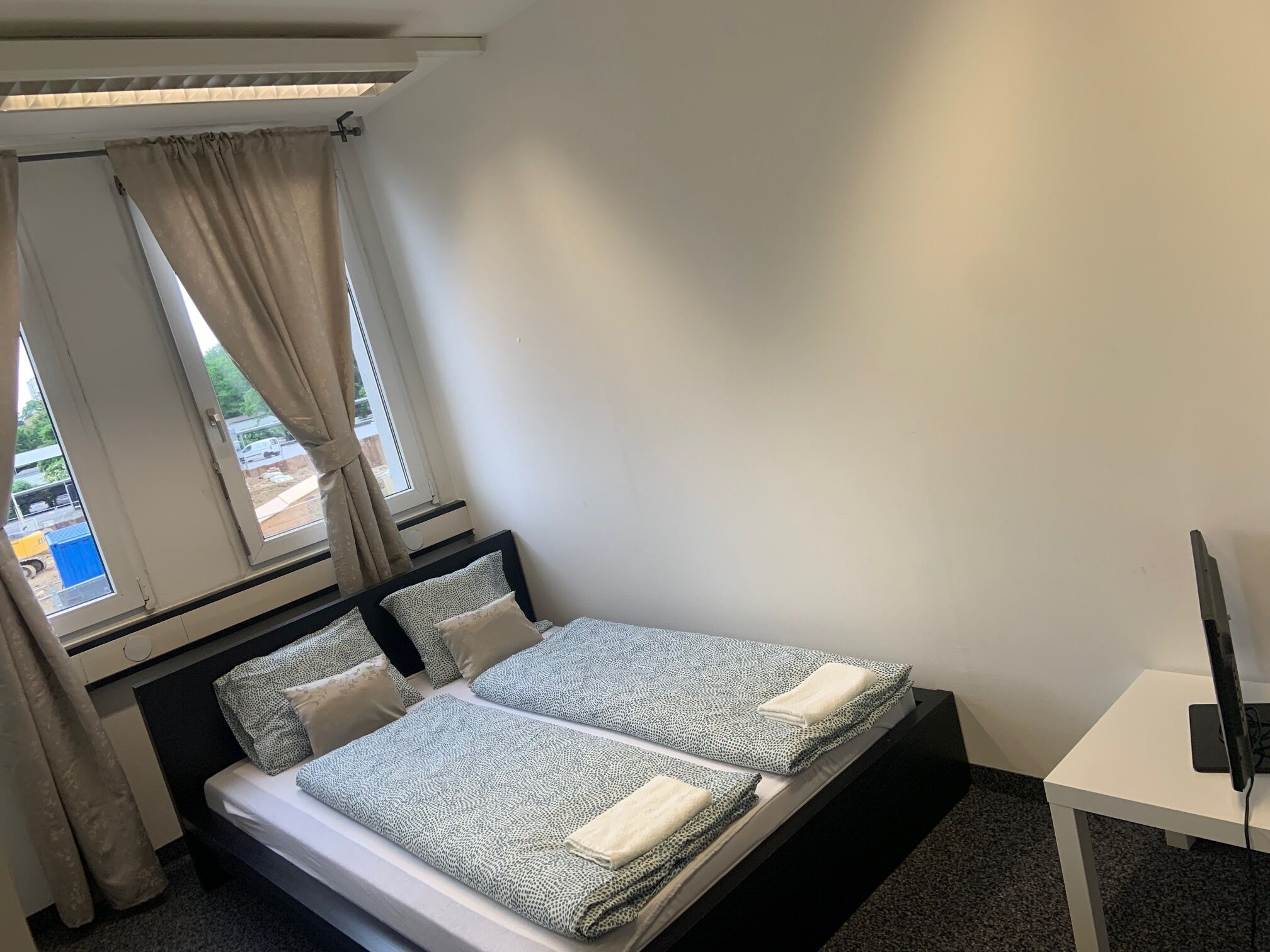 A single bed with a patterned bedspread, a bedside table with a flat-screen TV, and a window with curtains.