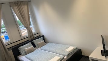 A single bed with a patterned bedspread, a bedside table with a flat-screen TV, and a window with curtains.