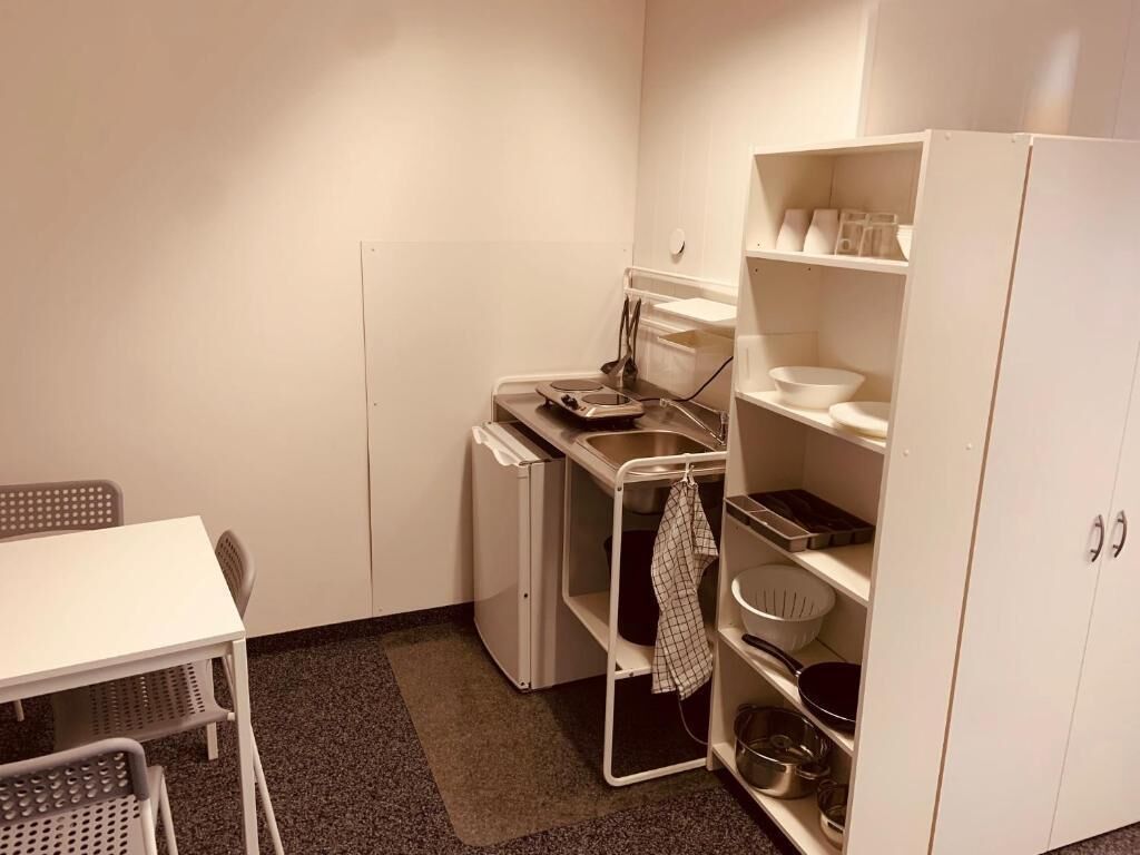 A compact kitchen with a white table, chairs, a small refrigerator, a sink, and a shelf with kitchenware.