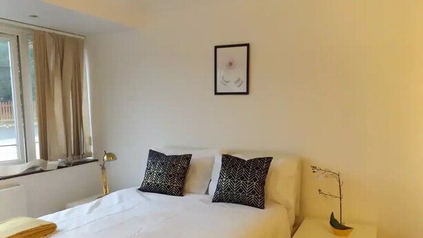 A neatly made bed with white bedding and patterned pillows, a bedside lamp, and a small table with a potted plant. - Coventry Park House (Coventry)