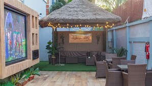 An outdoor patio with wicker furniture, a thatched-roof structure, and a flat-screen TV mounted on a brick wall. - Ivy Hotel Emerald (Lagos)