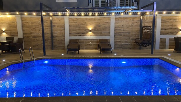 A swimming pool with a blue mosaic tile design, surrounded by lounge chairs and a wooden wall with built-in lighting. - Ivy Hotel Emerald (Lagos)
