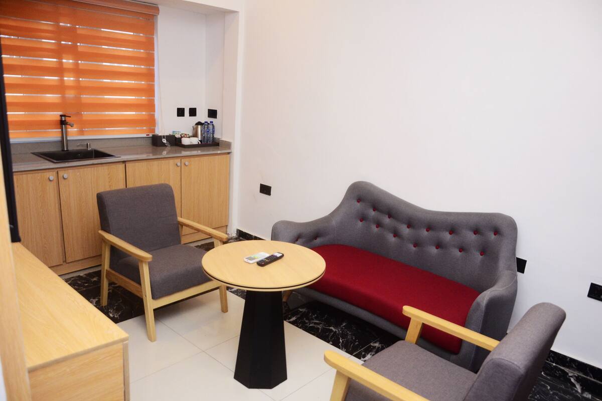A compact living space with a sofa, two armchairs, a small round table, a kitchen area with a sink and countertop, and a window with blinds.