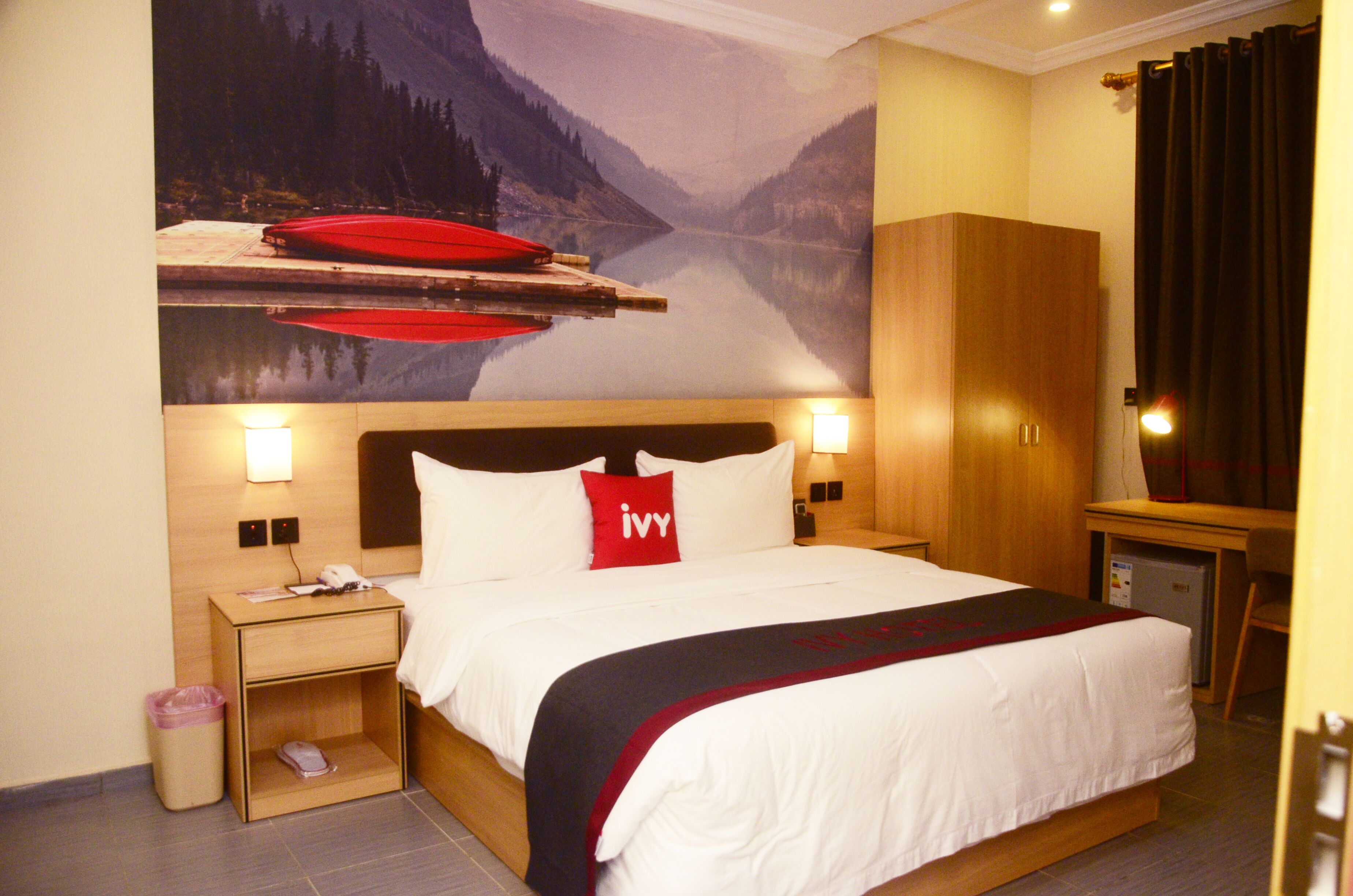 A hotel room with a bed, nightstands, a desk, and a view of a mountain lake.