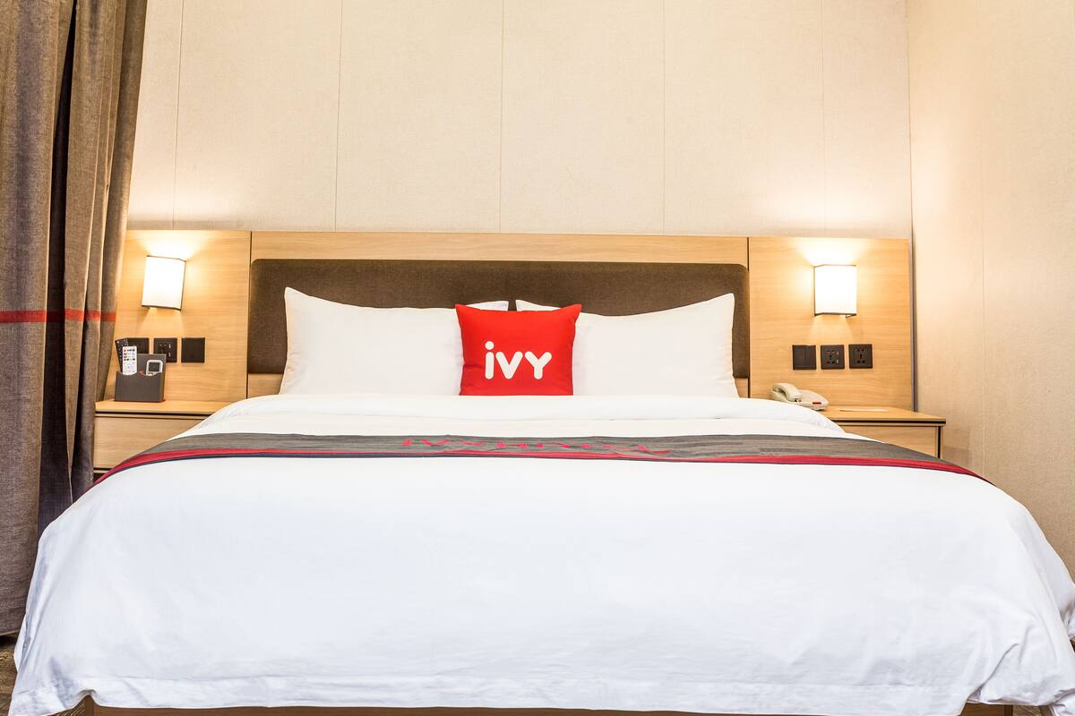 A hotel room with a large bed, a red pillow with 'Ivy' on it, two bedside tables with lamps, and a wooden headboard.