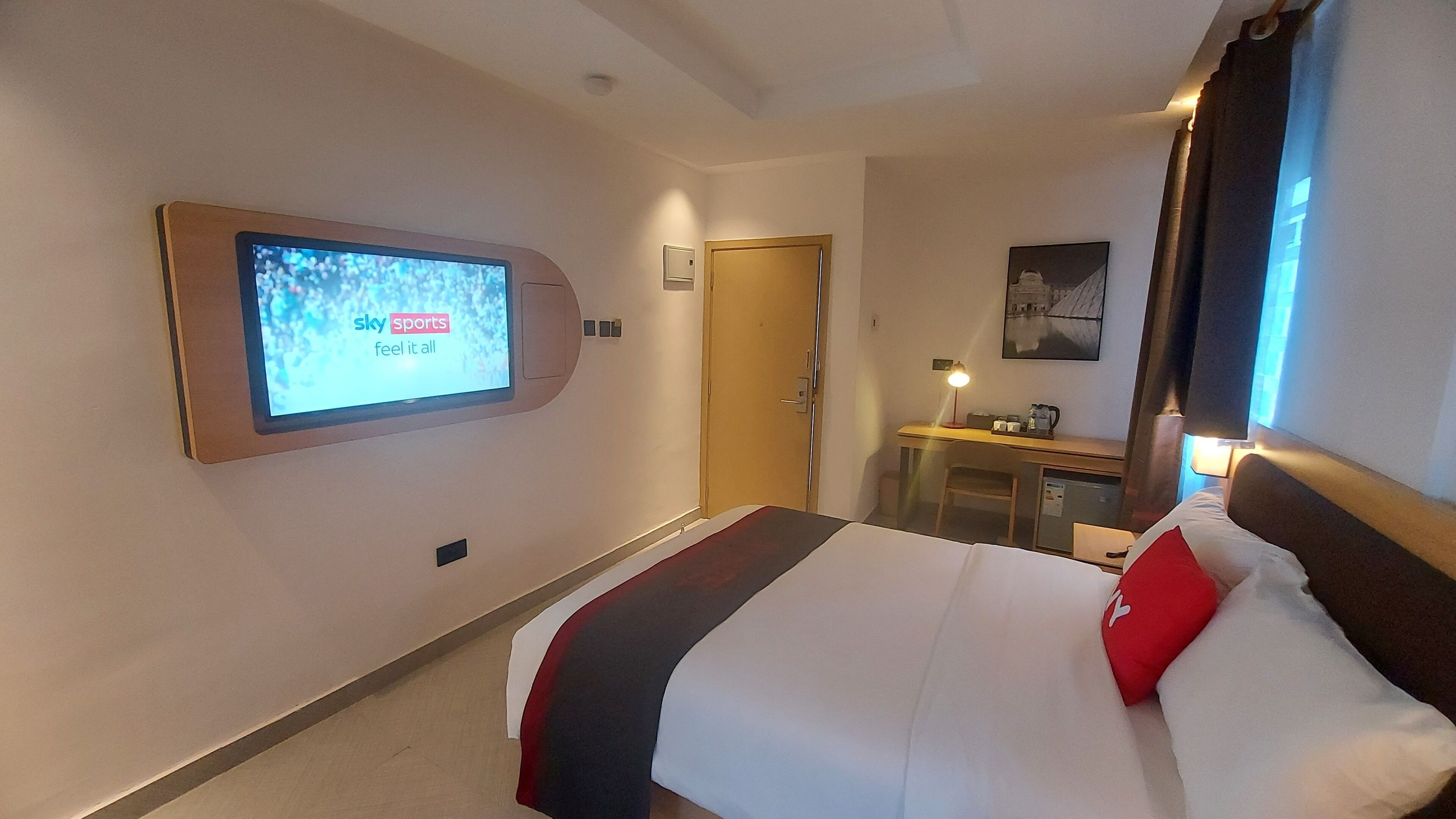 A hotel room with a bed, a desk with a lamp, a television mounted on the wall, and a door.