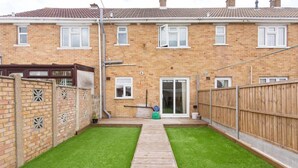 A residential backyard with a wooden deck, green artificial grass, and a brick house. - Immaculate 2-bed House in Dartford (Dartford)