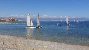 Un paysage côtier avec des voiliers sur une eau calme, une plage de galets et des montagnes lointaines sous un ciel dégagé.