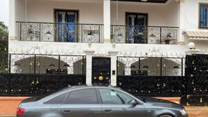 Een grijze auto geparkeerd voor een huis met sierlijke zwarte poorten en een balkon met decoratieve hekwerk.