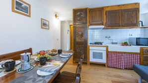Un coin repas avec une table en bois mise pour deux personnes, une machine à café et un panier de fruits. À côté se trouve une cuisine avec des placards en bois, un four à micro-ondes et une cuisinière.