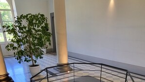 A hexagonal fire pit with a metal grate, surrounded by a black railing, in a spacious indoor area with a checkered floor and a large potted plant in the background.