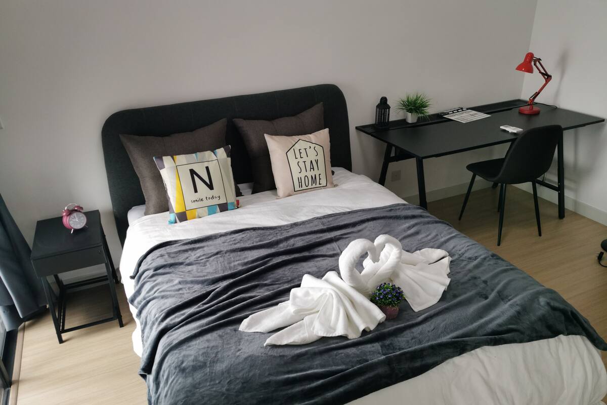 A neatly made bed with a grey and white duvet, two pillows with motivational quotes, a red alarm clock, a small potted plant, a desk with a red lamp, and a black chair.