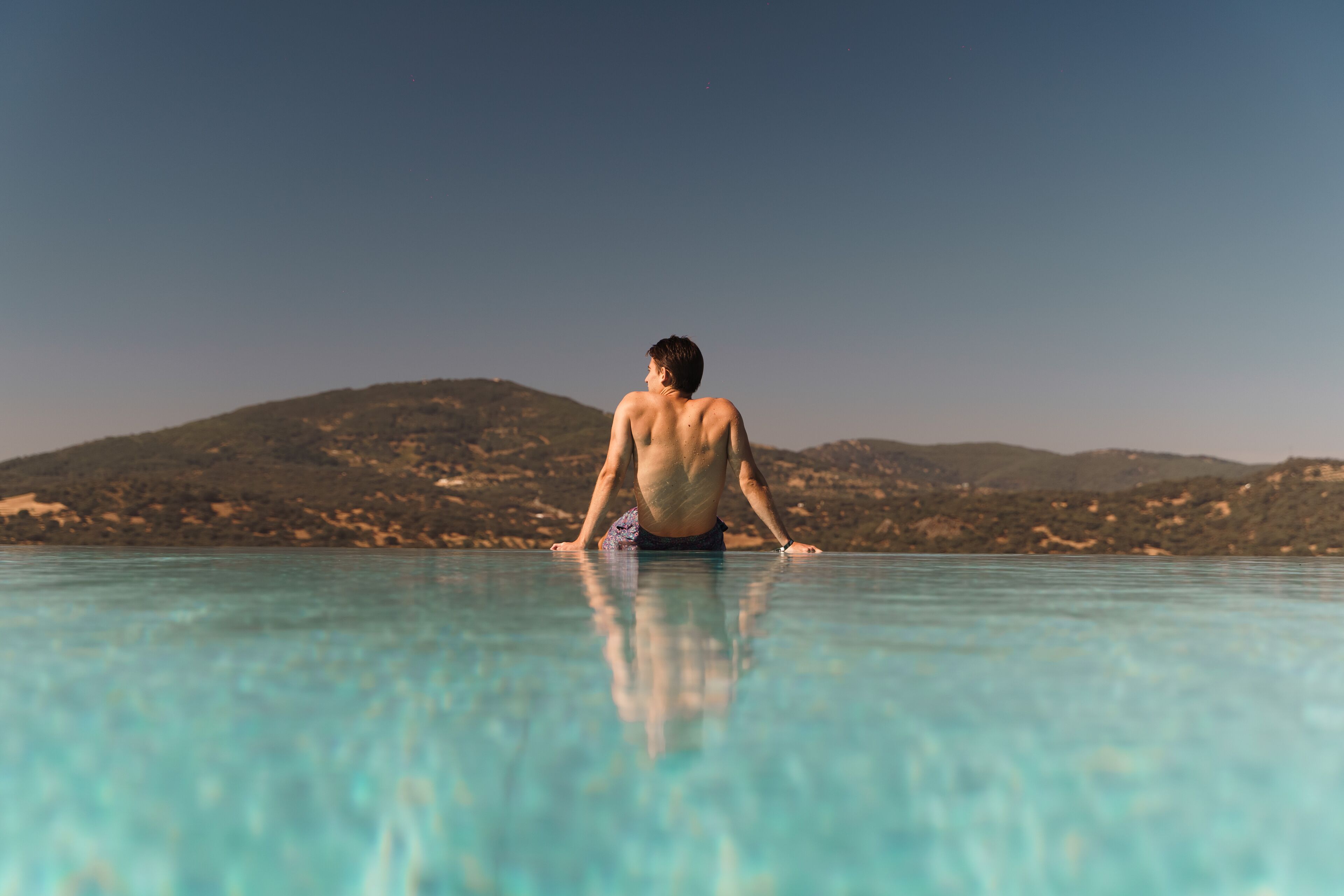 A person standing at the edge of a clear pool with a mountainous landscape in the background.
