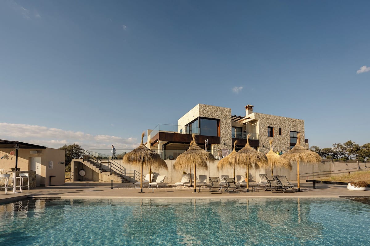 Un hotel moderno con piscina, sombrillas de paja y un cielo azul despejado.