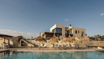 A modern hotel with a swimming pool, thatched umbrellas, and a clear blue sky.