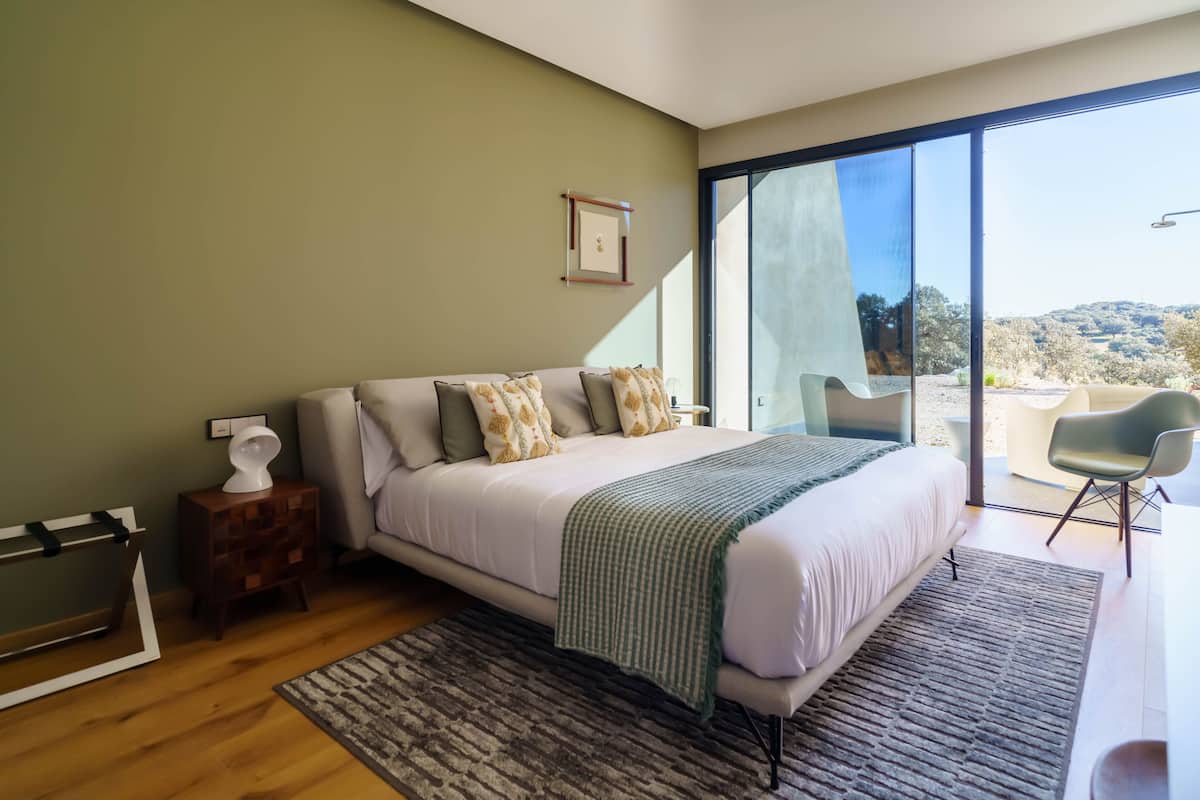Un dormitorio moderno con una cama grande, una mesita de noche, una silla y vistas al exterior a través de un ventanal corredero.
