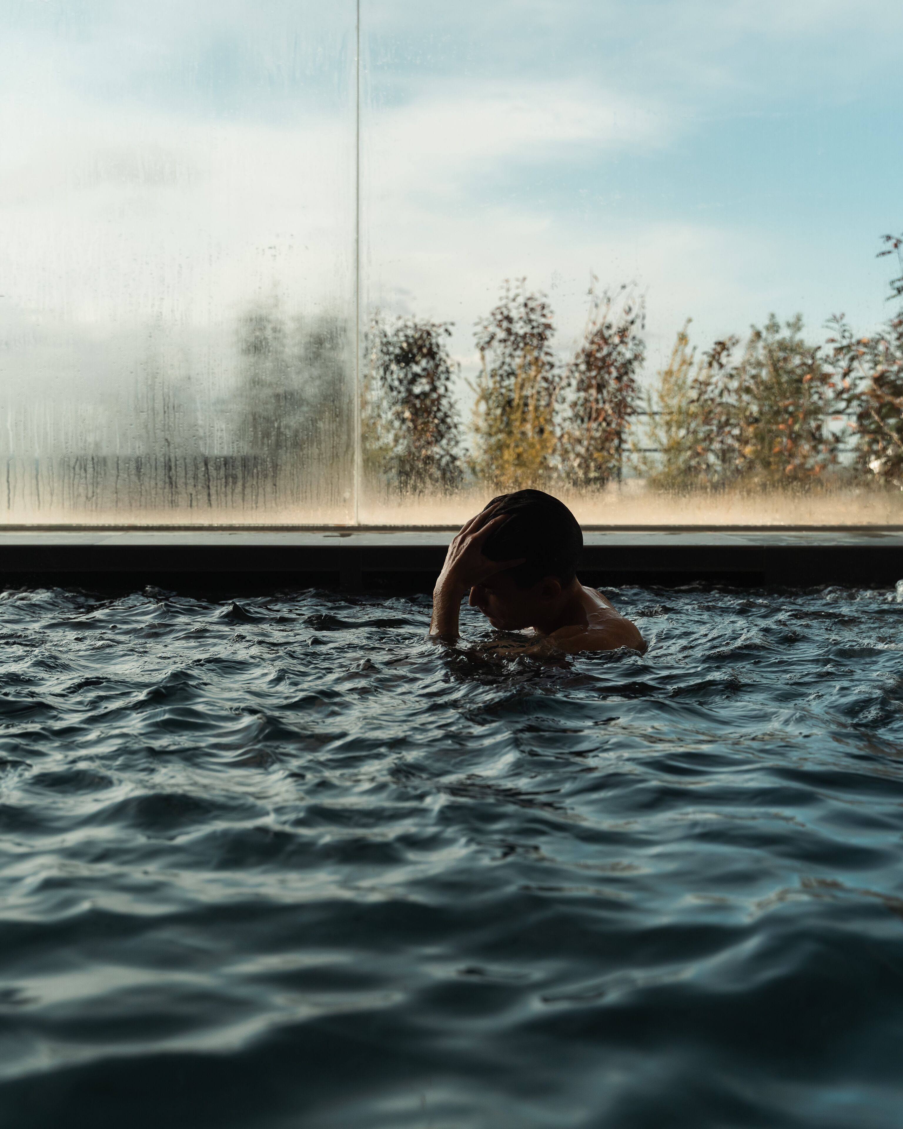 Une personne nage dans une piscine, avec des arbres visibles en arrière-plan et une fenêtre à l’horizon.