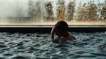 Une personne nage dans une piscine, avec des arbres visibles en arrière-plan et une fenêtre à l’horizon.