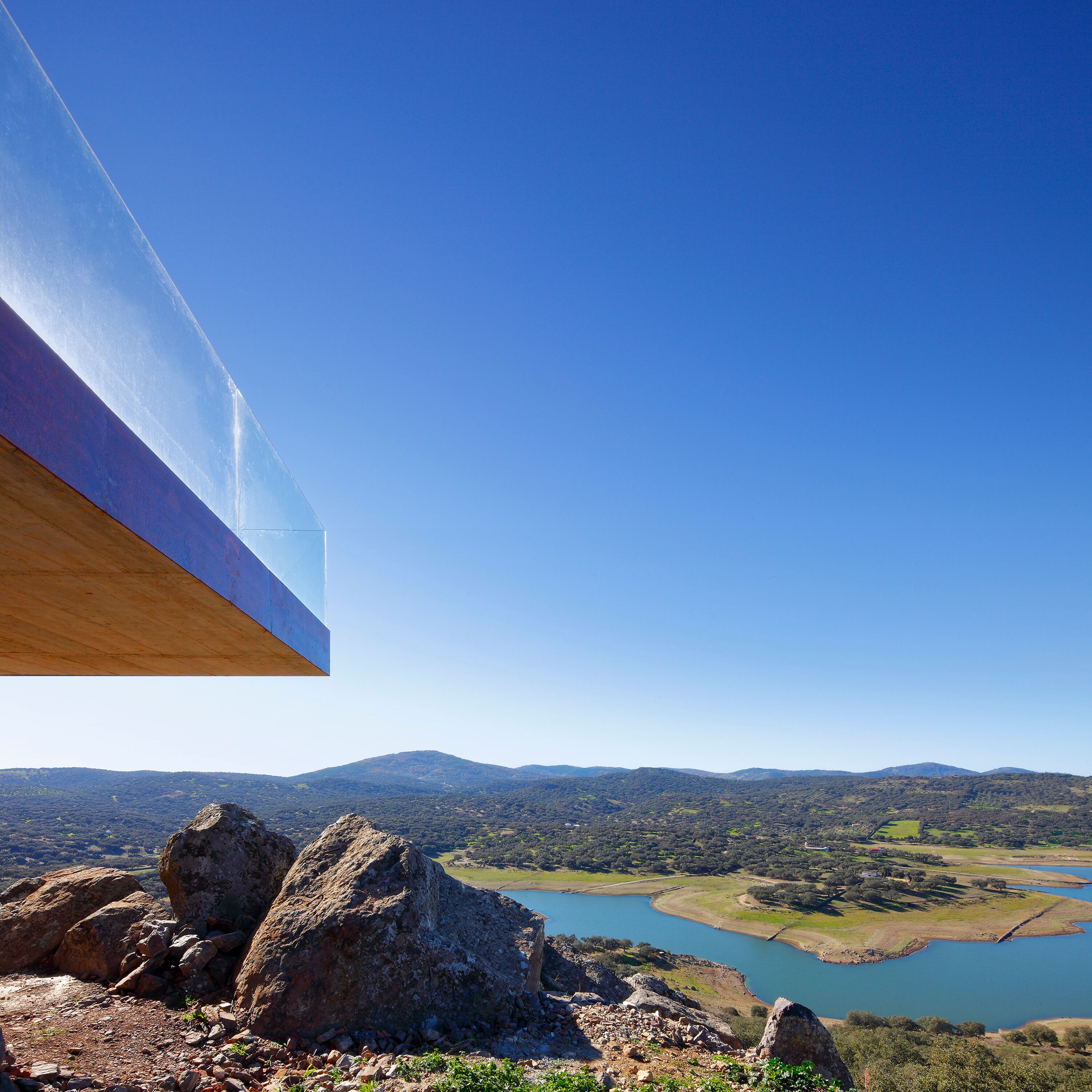 A modern building with a large glass facade overlooking a scenic landscape with mountains and a lake.