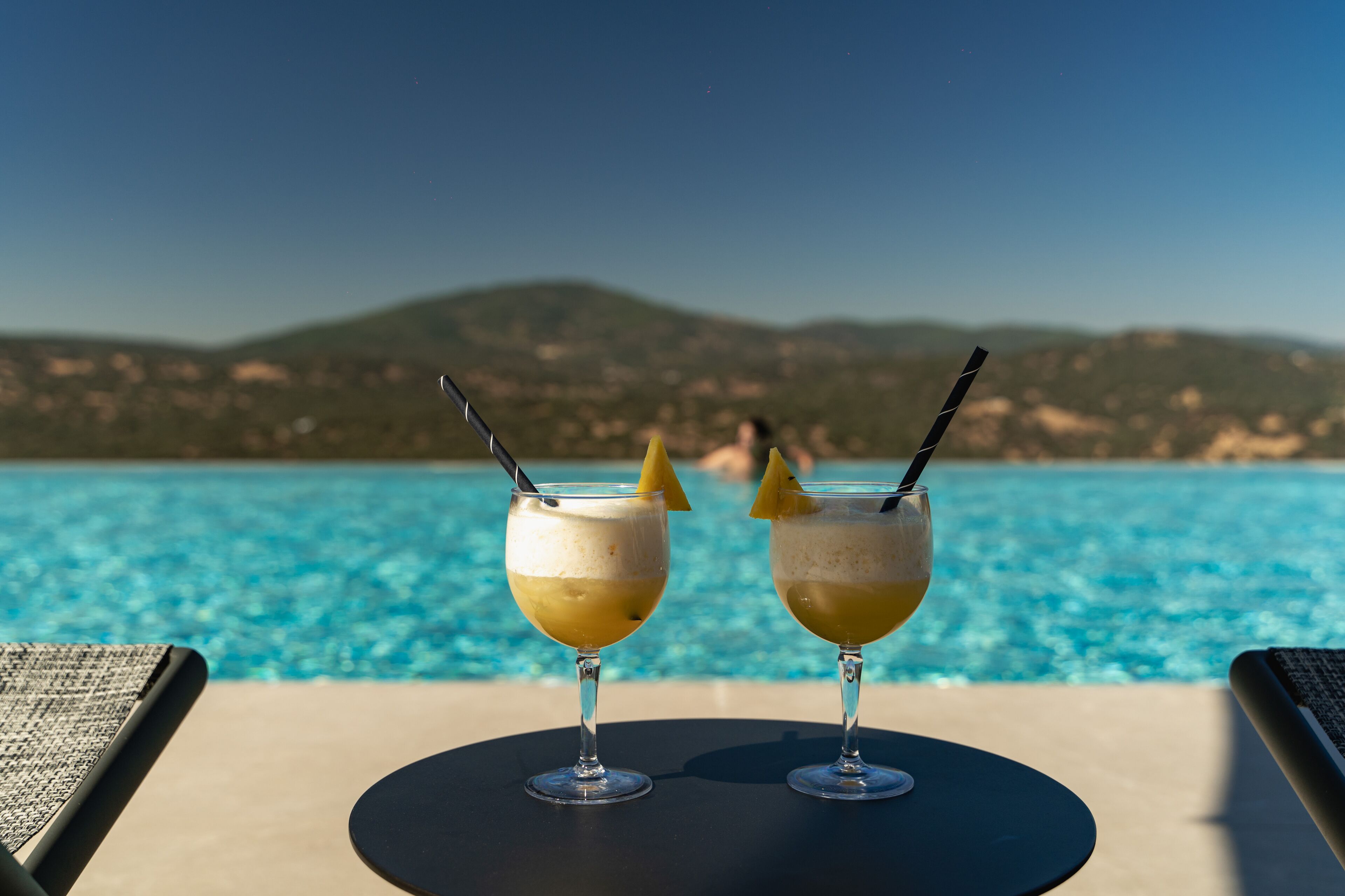 Zwei Cocktails auf einem Tisch neben einem Pool mit Blick auf die Berge.