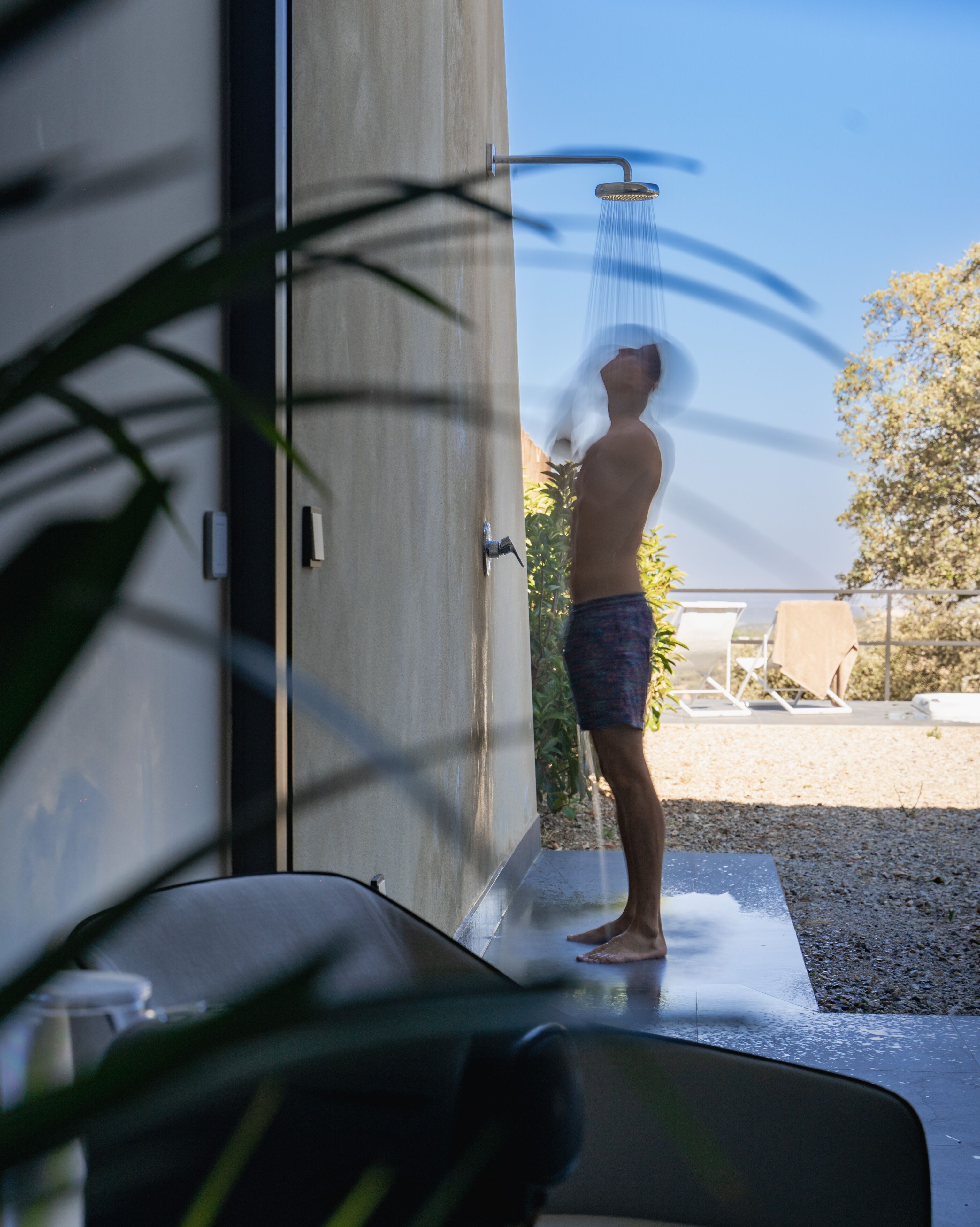 A person standing under a shower in a modern bathroom with a glass door, a plant in the foreground, and outdoor furniture visible through the door.