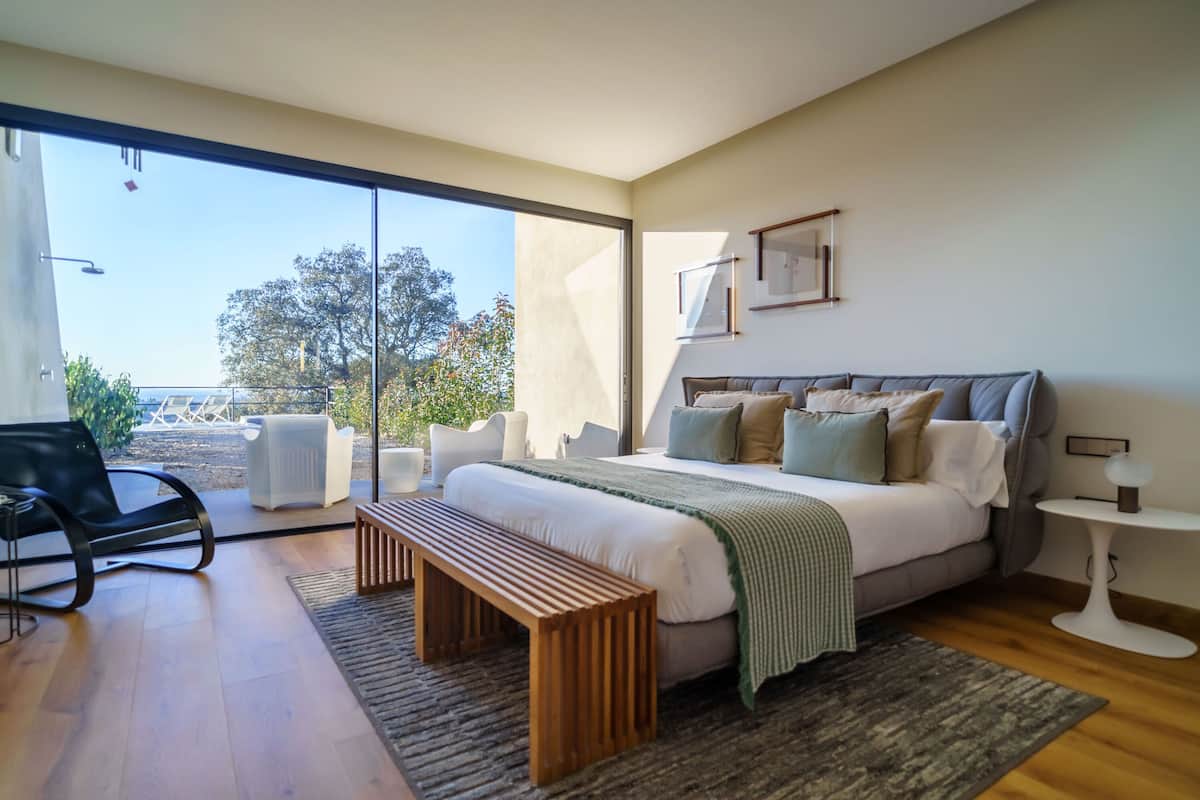 Un dormitorio moderno con una cama grande, un banco, una mesita y vistas al exterior.