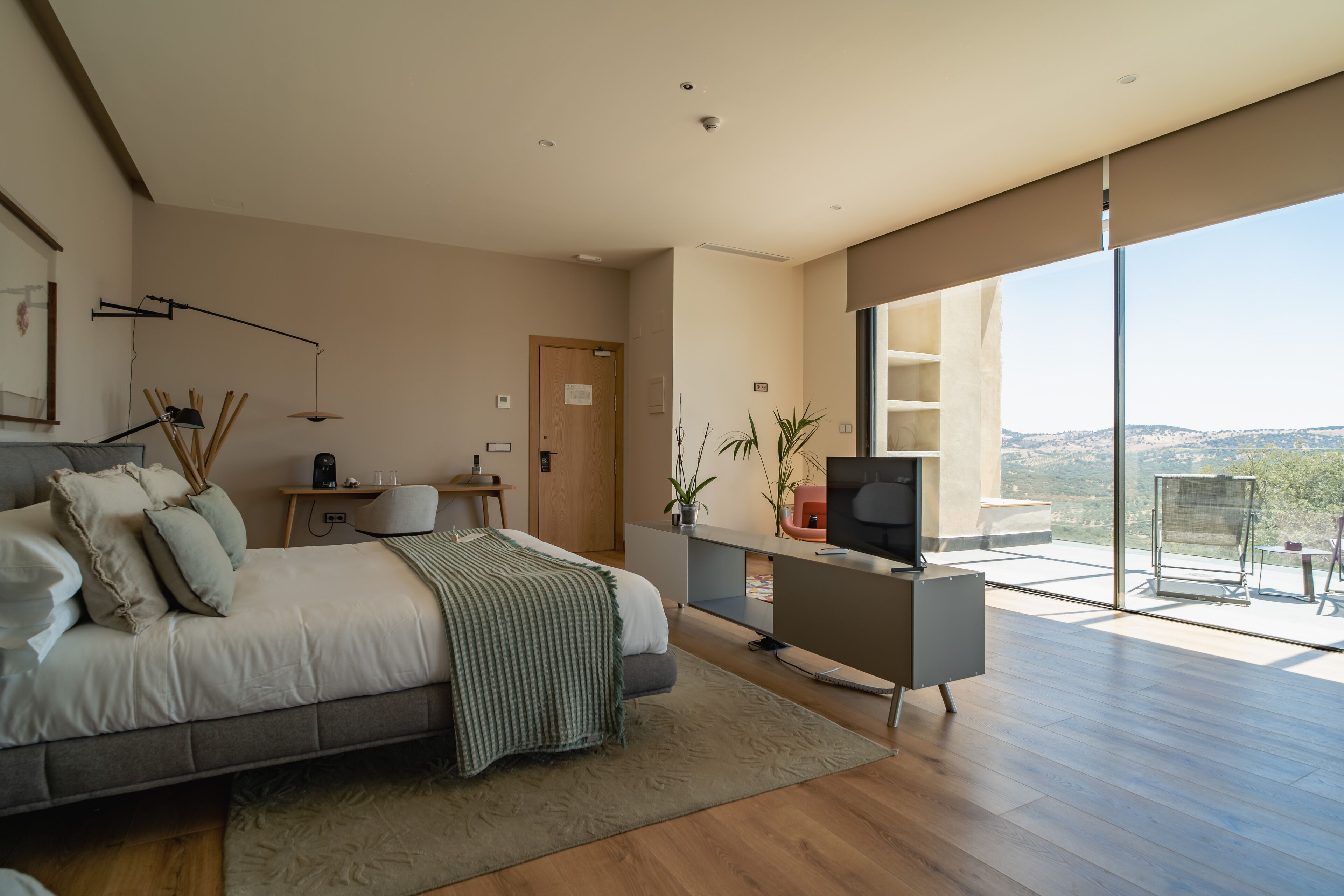 A modern bedroom with a large bed, a desk, and a TV. There is a balcony with a view of the landscape.