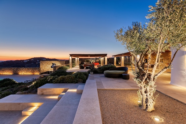 A modern outdoor patio with a stone wall, seating areas, and a view of the sea at dusk.