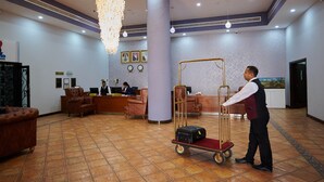 Un vestíbulo con mostrador de recepción, un pilar grande, una araña y un carrito de equipaje.