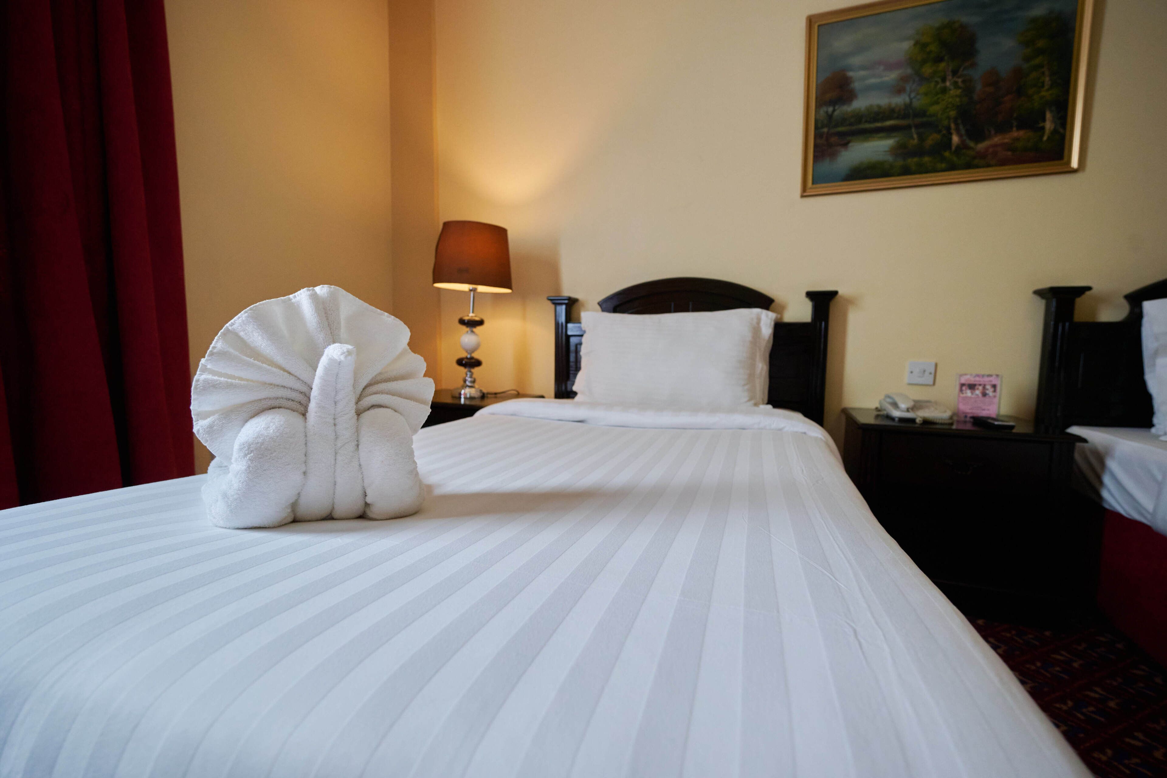 A hotel room with a neatly made bed, a lamp, a painting on the wall, and a red curtain.