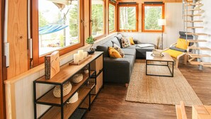 A cozy living room with a sofa, a coffee table, and a bookshelf. There are large windows with a view of greenery outside.