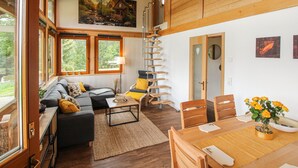 A spacious living room with a sofa, a dining table, and a chair. There is a painting on the wall and a window with a view of trees.