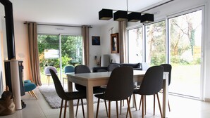 A modern dining area with a large table and chairs, a fireplace, and large windows offering a view of the outdoors. - Maison, 5mn de Plage Lanséria, 4 à 10 Personnes (Mesquer)