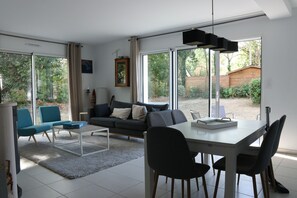 Relax in a spacious living area with a central seating space, dining table, and large windows offering views of a lush garden.