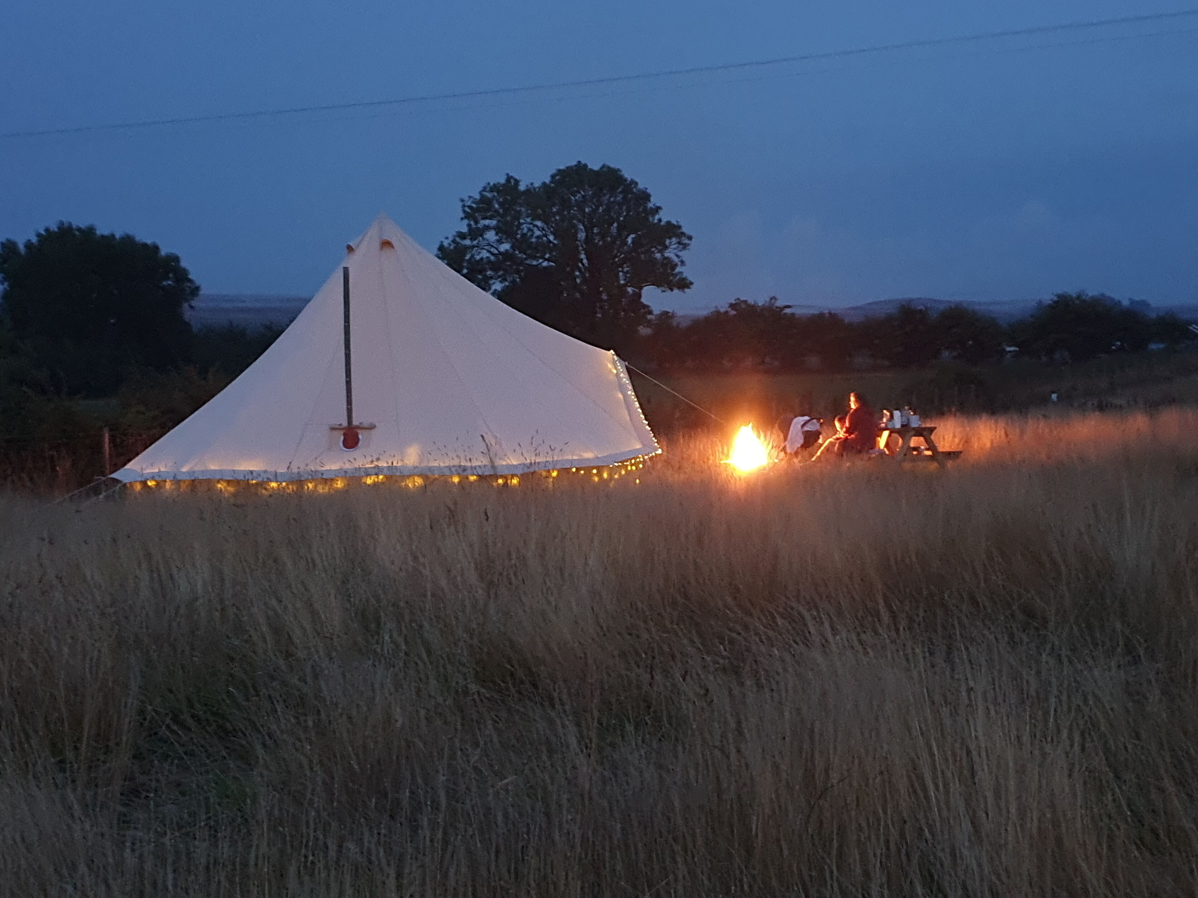 A white tent with string lights, set in a grassy field with a fire in the background.