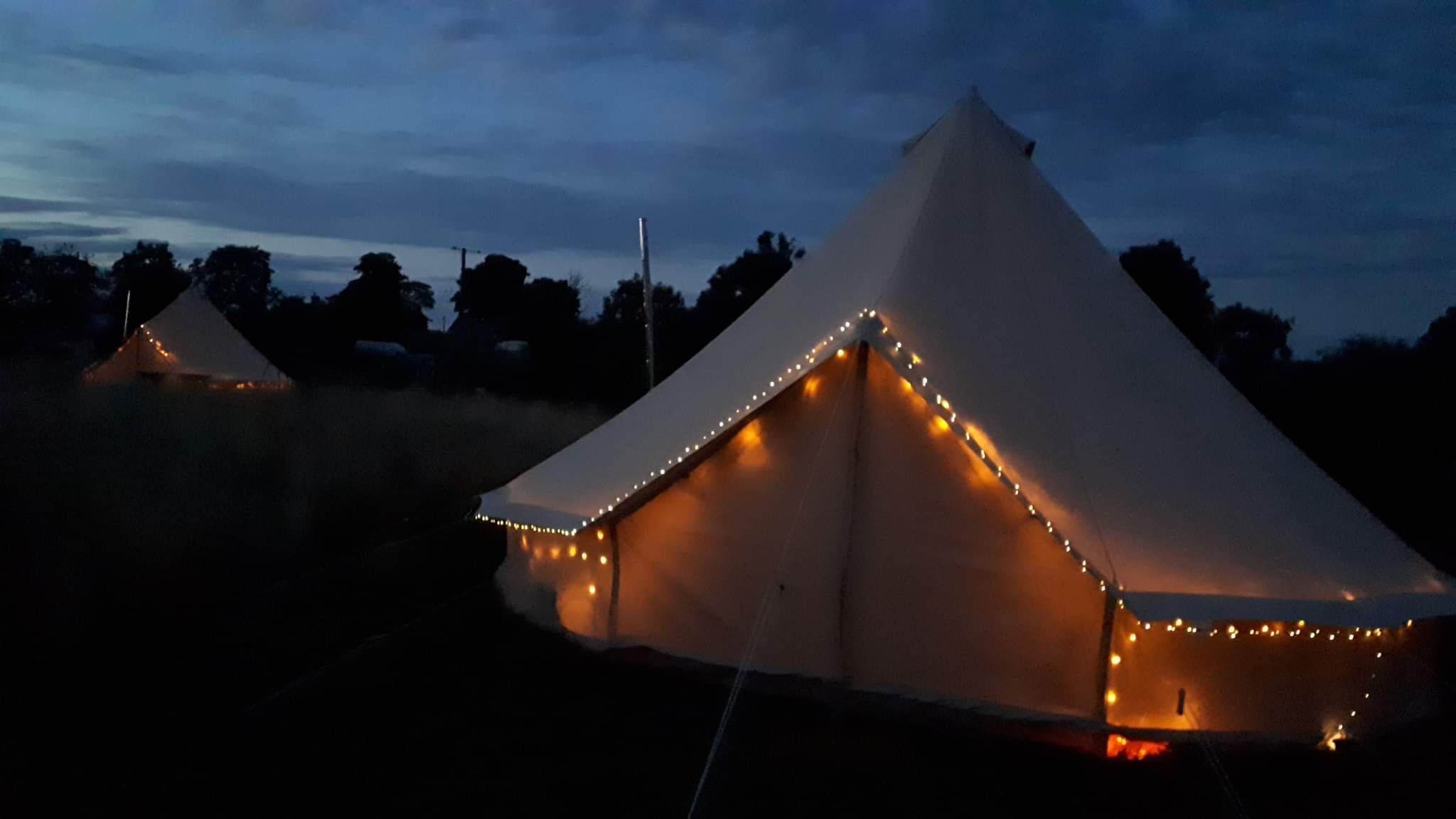 A series of white tents with string lights along the edges, set in a natural landscape at dusk.
