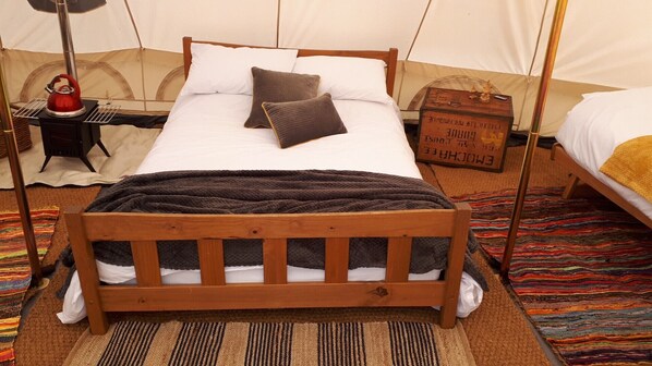 Una cama acogedora con estructura de madera, ropa de cama blanca y un plaid oscuro. Hay una mesita con una tetera y un letrero al lado de la cama.