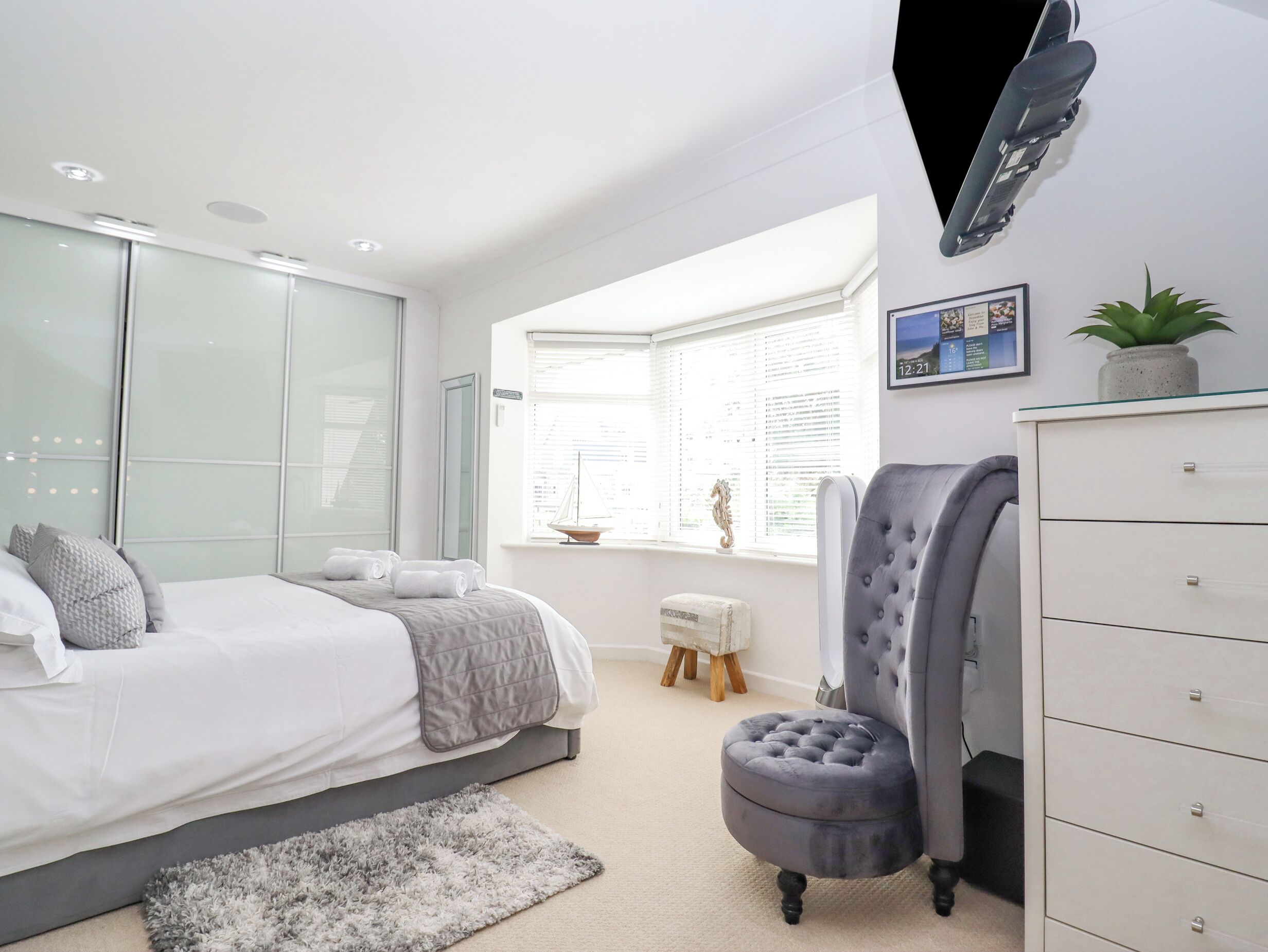 A modern bedroom with a bed, a grey upholstered chair, a white dresser, and a window with blinds.