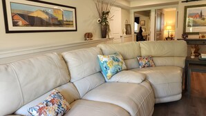 Una sala de estar con un sofá seccional de cuero color beige, dos almohadas decorativas con motivos florales, un cuadro de paisaje enmarco y un jarrón con plantas secas.