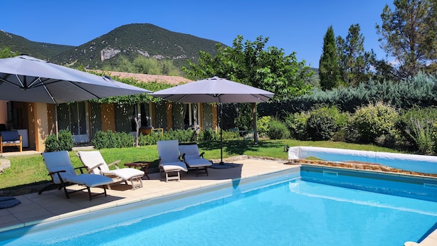 Zona de piscina con sillas de descanso y sombrillas, rodeada de vegetación y una montaña al fondo.
