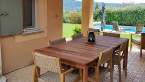 Un patio al aire libre con mesa y sillas de madera, vistas a una piscina y una zona cubierta con techo de madera.